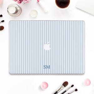 May include: A light blue and white striped laptop case with the Apple logo and the initials "SM" in blue. The case is surrounded by makeup brushes, a cup of coffee, and beauty products.