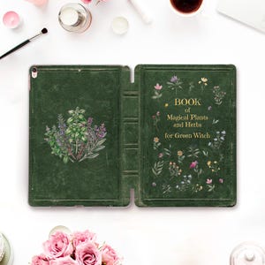 May include: A dark green tablet case featuring a textured leather-like finish. The front cover displays the gold-lettered text "BOOK of Magical Plants and Herbs for Green Witch" surrounded by small floral illustrations, while the back shows herbs.