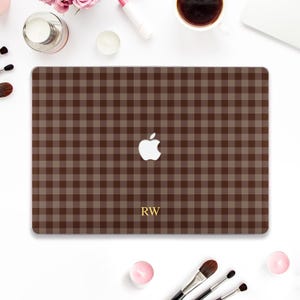 May include: A rectangular laptop skin featuring a brown and tan gingham plaid pattern. The center displays a white Apple logo cutout, and the letters RW are printed in a gold serif font near the bottom edge of the laptop cover surface.