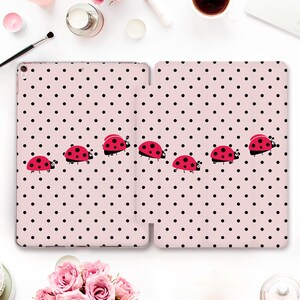 Może przedstawiać: Etui na tablet z jasnoróżowym tłem, ozdobione powtarzającym się wzorem małych czarnych kropek oraz kilkoma czerwonymi biedronkami z czarnymi kropkami i białymi oczami, ułożonymi w poziomą linię na powierzchni pokrowca.