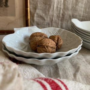 May include: A stack of white, leaf-shaped ceramic dishes with three walnuts in the center. The dishes have a scalloped edge and a subtle embossed design. A stack of similar dishes is in the background, and a framed picture is visible.