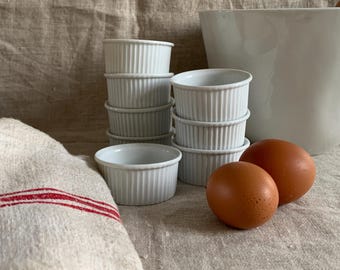 以下が含まれることがあります： 白く溝の入ったラメキンがグループで積み重ねられ、茶色の卵2個と大きな白いミキシングボウルが並んでいます。赤いストライプの入ったリネンのタオルが前景にあります。ベーキングや料理を連想させるシーンです。