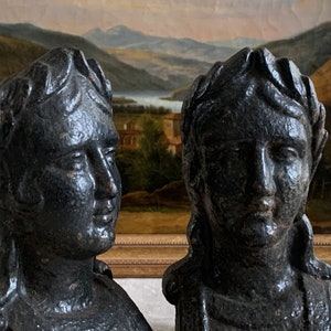 Puede incluir: Se muestran dos bustos grises oscuros de figuras clásicas, posiblemente romanas o griegas. Los bustos presentan rasgos faciales detallados y están adornados con coronas de laurel. El fondo incluye una pintura de paisaje con un lago y montañas.
