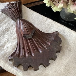 Puede incluir: Una pieza decorativa de madera tallada, de color marrón oscuro, con un diseño floral. La pieza tiene una superficie detallada y texturizada y un borde dentado. Descansa sobre una tela color crema.