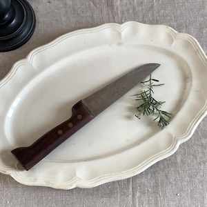 Op de afbeelding: Een ovale, crèmekleurige serveerschaal met een geschulpte rand. Een vintage mes met een donkerhouten handvat en een zilveren lemmet ligt op de schaal, samen met een takje rozemarijn. Een zwarte kandelaarvoet staat op de achtergrond.