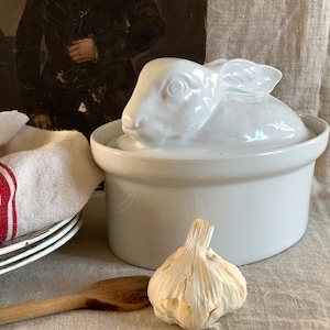 May include: A white ceramic lidded tureen or serving dish featuring a sculpted rabbit figure on the lid. The container has a smooth, glossy finish and an oval shape. The lid is designed with the rabbit resting on top, serving as a handle.