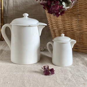 Può includere: Due teiere in ceramica bianca, una grande e una piccola, con coperchi e beccucci, sono appoggiate su una superficie neutra. La teiera più grande ha un manico curvo. Un fiore secco è posato vicino. Un cesto di vimini e un'opera d'arte incorniciata sono sullo sfondo.