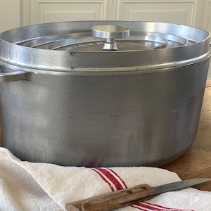 Può includere: Una pentola olandese grande in alluminio argentato con coperchio. La pentola ha un fondo arrotondato e un bordo largo e piatto. Il coperchio ha un piccolo pomo rotondo in cima.