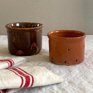 Puede incluir: Dos macetas cilíndricas de cerámica con pequeñas perforaciones circulares en los lados. Una tiene un esmalte marrón oscuro brillante, mientras que la otra presenta un acabado mate en terracota. Ambas tienen un borde sencillo y una base plana.