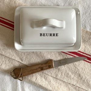 Könnte beinhalten: Eine weiße, rechteckige Butterdose aus Keramik mit einem Deckel, der einen Griff und das in Schwarz aufgedruckte Wort BEURRE aufweist. Unter der Dose liegt ein kleines Messer mit Holzgriff und Metallklinge sowie einer Lederschlaufe.