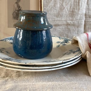 Könnte beinhalten: Ein blaues Keramikgefäß mit Deckel steht auf einem Stapel weißer Teller mit blauen Blumenmustern. Ein Leinentuch mit roten Streifen befindet sich rechts. Das Gefäß hat ein rustikales Aussehen.