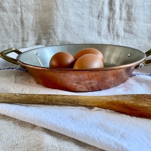 May include: A copper pan with brass handles containing three brown eggs, set on a white cloth. A wooden spoon lies in front. The pan's interior is silver. The eggs are a staple of British cuisine.