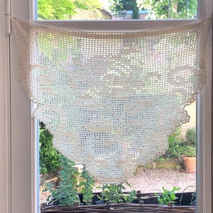 May include: Cream-colored, crocheted window valance with a scalloped edge. The delicate lace curtain hangs in front of a window, with a view of greenery and a small flower pot. The valance is made of a fine, open weave.