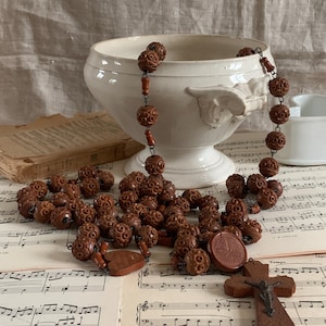 以下が含まれることがあります： 精巧に彫刻された茶色のビーズとサンゴ色のスペーサーが付いた、ヴィンテージのロザリオ。ロザリオは白いセラミックボウルにかけられ、楽譜の上に置かれています。木製の十字架やその他の宗教的なアイテムも存在します。