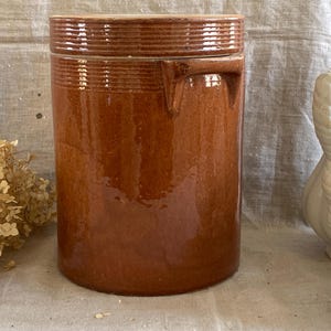 Puede incluir: Un tarro de cerámica esmaltada marrón con tapa y asa. El tarro tiene un aspecto rústico y es perfecto para guardar productos secos.