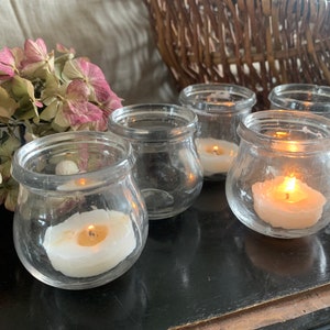Pode incluir: Uma coleção de pequenos castiçais de vidro transparente. Alguns contêm velas brancas acesas, emitindo um brilho quente. Os castiçais estão dispostos sobre uma superfície escura, com flores secas e uma cesta de vime ao fundo.