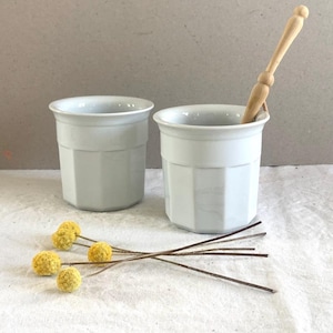Könnte beinhalten: Zwei weiße Keramikbecher mit einem facettierten, mehrseitigen Außendesign. Ein Becher enthält einen hellen Rührstab aus Holz. Vier getrocknete gelbe Trommelstöckchen-Blüten mit dünnen braunen Stielen sind vor den Bechern angeordnet.