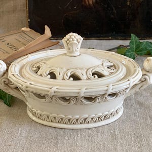 Pode incluir: Tigela com tampa de cerâmica branca suja com detalhes ornamentados. A tampa apresenta um botão e um design vazado. A tigela tem alças com rostos esculpidos. Estilo vintage, adequado para exibição ou armazenamento.