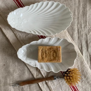 May include: Two white, shell-shaped ceramic dishes, one holding a bar of soap, and a wooden-handled brush rest on a linen cloth with red stripes. The dishes have a scalloped edge and embossed details.