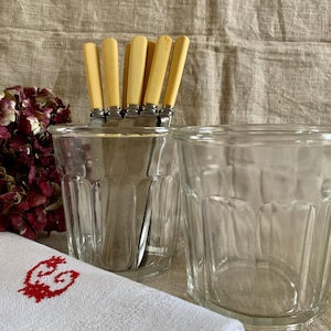 May include: Clear, faceted glass tumblers with a set of knives with cream-coloured handles. A white linen napkin with a red embroidered design is in the foreground. Dried burgundy flowers are visible on the left.