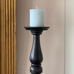 Può includere: Una candela a colonna azzurra è posta su un candeliere in legno nero. Il candeliere presenta un design tornito e una base rotonda. Lo sfondo include uno specchio con cornice dorata e una parete colorata neutra, creando un'estetica classica ed elegante.