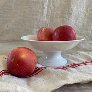 Puede incluir: Un frutero blanco con pedestal contiene dos manzanas rojas, con otra manzana sobre un paño a rayas. Las manzanas son de un rojo vibrante, y el frutero tiene un diseño clásico. El paño tiene rayas rojas.