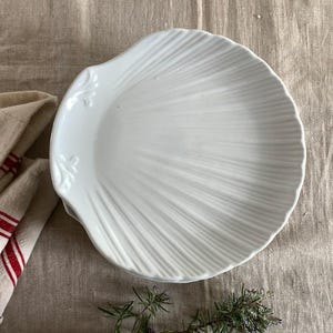 May include: A white, shell-shaped plate with a ridged design. The plate has a scalloped edge and decorative floral accents. The plate is on a neutral-colored linen surface, with a folded linen napkin with red stripes and sprigs of rosemary.