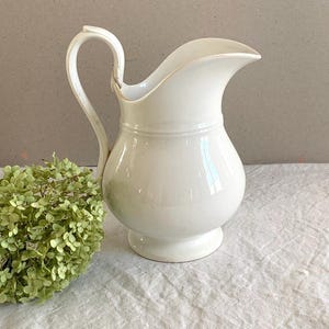 Pode incluir: Um jarro de cerâmica branca com asa curva, bico largo e corpo arredondado, posicionado ao lado de um cacho de flores de hortênsia secas verde-claras. O jarro apresenta um acabamento liso e brilhante com um detalhe de faixa horizontal.