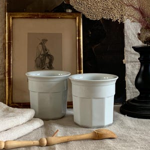 Può includere: Due vasi in ceramica bianca con un design geometrico. I vasi sono cilindrici con un bordo leggermente svasato. Un cucchiaio di legno e una pila di asciugamani sono in primo piano. Un quadro incorniciato e un candelabro nero sono sullo sfondo.