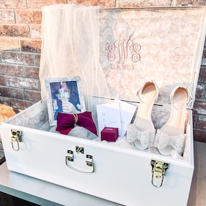 May include: White wooden keepsake box with a gold latch and handle. The box is open and contains a photo, a burgundy bow, two white shoes with bows, and a small red ring box. The inside of the lid is lined with a light pink marble pattern and has the text "J.J.S. 12.8.2024" in gold lettering.