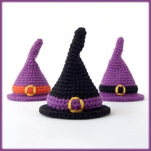 Può includere: Tre cappelli da strega all'uncinetto in viola e nero, ciascuno con una fascia a contrasto e una fibbia color oro. I cappelli sono disposti su una superficie bianca, perfetti per Halloween.