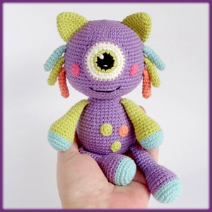 Puede incluir: Un juguete monstruo de crochet morado con un solo ojo grande, detalles verdes y naranjas y una expresión amigable.