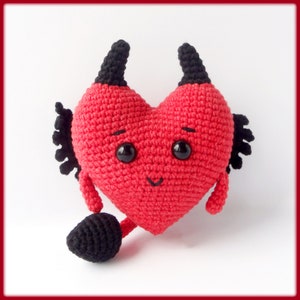 May include: A red crocheted heart-shaped plush toy with black horns, a tail, and a smiling face.