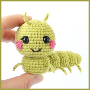 May include: A handmade, crocheted caterpillar toy in shades of green. The caterpillar has a round head with black eyes, pink cheeks, and a smiling mouth. It has small antennae and is held by a hand.