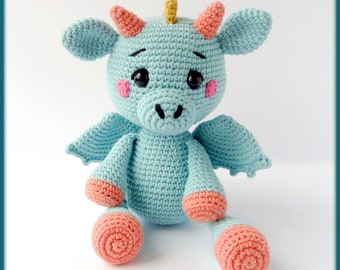 May include: A light blue crocheted dragon with pink cheeks, a yellow horn, and pink feet. The dragon is sitting with its wings spread out.