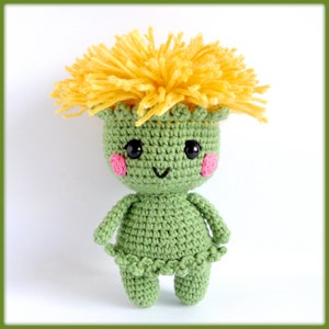 May include: A green crocheted plush toy with a yellow crocheted flower on top. The toy has a smiling face with black eyes and pink cheeks.