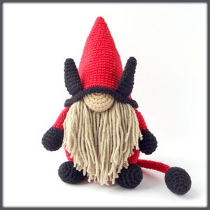May include: A red crocheted gnome with black horns, a black hat, and a long, white beard. The gnome has a black tail and is sitting on a white background.
