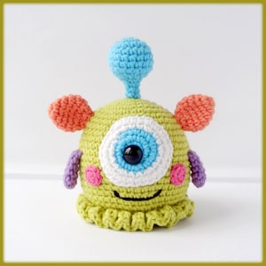 May include: A crocheted green monster toy with a single large eye, pink cheeks, and a blue antenna. The monster has orange ears and purple arms.