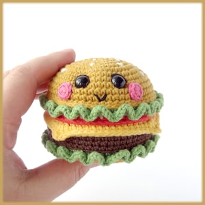 Può includere: Un giocattolo di peluche a forma di hamburger all'uncinetto con un viso sorridente, lattuga verde, pomodoro rosso, formaggio giallo e un hamburger marrone.