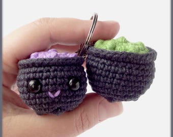 Puede incluir: Dos llaveros de caldero negro hechos a crochet con caras lindas. Un caldero está lleno de hilo morado y el otro de hilo verde.