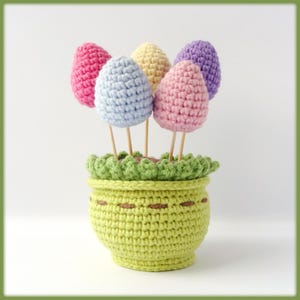 May include: A crocheted flower pot with five colorful crocheted eggs in a green pot. The eggs are pink, light blue, yellow, purple, and pink.