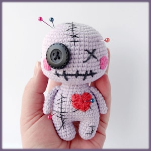 May include: A purple crocheted voodoo doll with a black button eye, pink cheeks, and a red heart on its chest. The doll has stitches on its face and body and is decorated with safety pins.