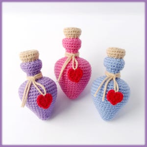 Puede incluir: Tres botellas de corazón de ganchillo con tapones de corcho y adornos de corazón rojo. Las botellas son en tonos de rosa, morado y azul.