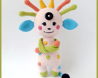 May include: A crocheted plush toy with a pink body, green arms, and a blue and orange head. The toy has a single black button eye and a black tail.