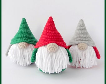 May include: Three handmade crochet gnomes with white beards and beige noses. Each gnome has a different colored hat: green, red, and gray. The gnomes are sitting on a white surface.