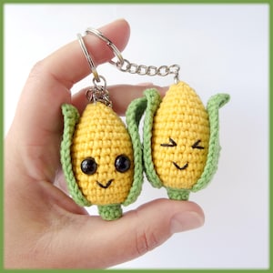 May include: Two crocheted corn keyrings. Each keyring features a yellow corn cob with green husks and black eyes, and a smiling mouth. The keyrings are attached to silver key rings.