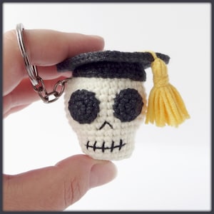 May include: A white crocheted skull wearing a black graduation cap with a yellow tassel. The skull has black eyes and a stitched mouth.