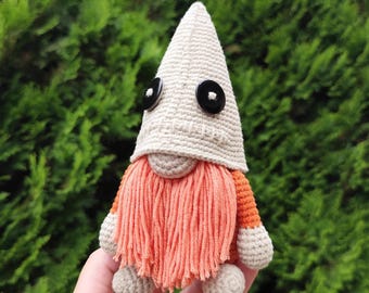 May include: A small, handmade crochet gnome figure featuring a tall, beige pointed hat with two black circular buttons for eyes. The gnome has a long, textured orange yarn beard, orange sleeves, and beige hands and feet, crafted with visible yarn stitches.