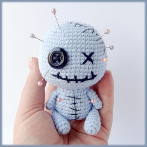 May include: A light blue crocheted voodoo doll with a black button eye, stitched mouth, and X for eyes. The doll is decorated with several pins with pink and silver beads.