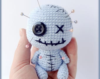 May include: A light blue crocheted voodoo doll with a black button eye, stitched mouth, and X for eyes. The doll is decorated with several pins with pink and silver beads.